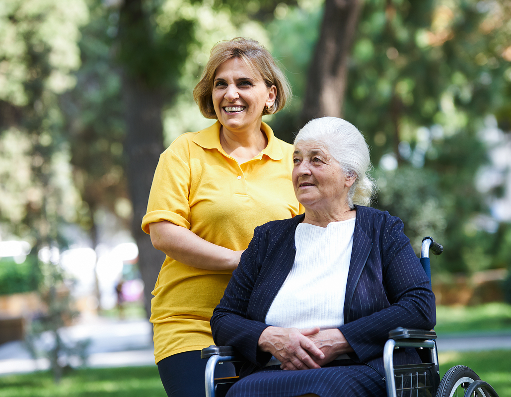 A smiling woman in a yellow shirt standing behind an elderly woman in a wheelchair outdoors, with trees and greenery in the background.
