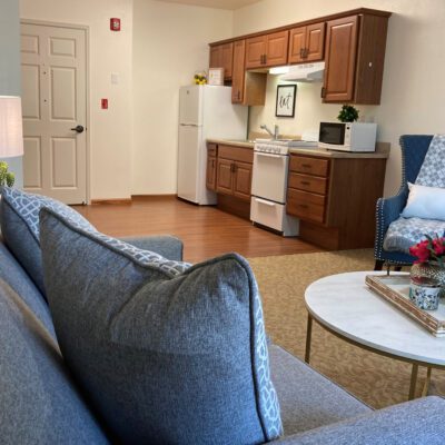 A cozy living area with a kitchen in the background, featuring wooden cabinets, a white refrigerator, stove, microwave, and small decorative items. In the foreground, there are blue upholstered chairs and a round coffee table with a tray and small flower arrangement.