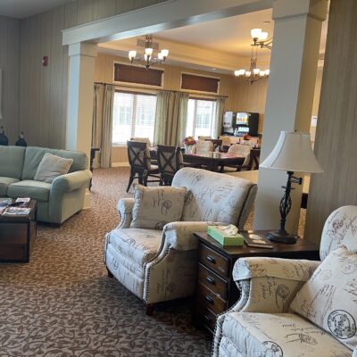 A spacious lounge area with upholstered armchairs, a sofa, and side tables, leading into a dining room with a large table and chairs, and multiple windows allowing natural light.