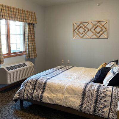 A cozy bedroom with a large bed covered in a patterned bedspread and decorative pillows. There is a window with checked curtains on the left and a wall-mounted wooden decorative piece above the bed. An air conditioning unit is below the window.