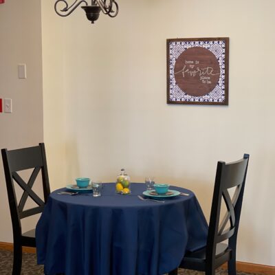 A round table covered with a blue tablecloth, set with four turquoise bowls, four glasses, and a small glass jar with a yellow lemon inside. Two black chairs with X-shaped backs are positioned around the table. A framed wall art piece with the words 