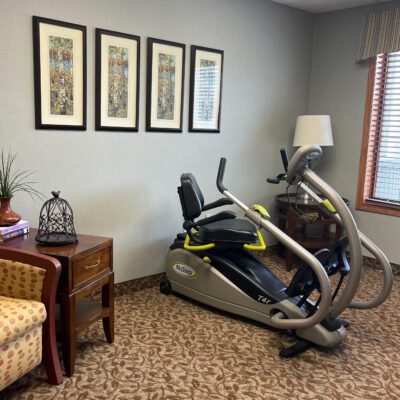 A room with four framed artworks on the wall, a wooden side table with decorative items, a patterned armchair, and a piece of exercise equipment, such as a recumbent bike, near a window with blinds.