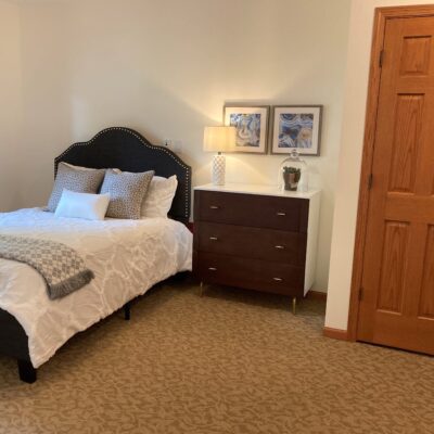 A bedroom with a bed featuring a black, tufted headboard, white bedding, and multiple pillows. Next to the bed is a dark wooden dresser with three drawers, topped with a lamp, framed artwork, and a small decorative object. There is a wooden closet door on the right side. The room has beige carpet and light-colored walls.