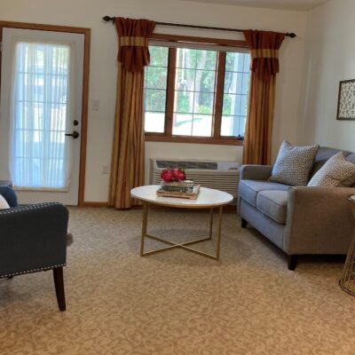 A cozy living room with a beige carpet, a gray sofa with patterned cushions, a round coffee table with a floral arrangement, a window with gold and rust-colored curtains, and a glass-paneled door with a sheer curtain.