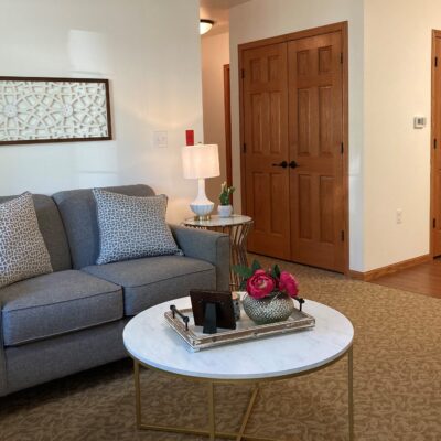 A cozy living room corner with a gray sofa adorned with patterned pillows, a round white coffee table with a decorative tray, a vase of pink flowers, and a picture frame. There is a wooden door, a wall-mounted frame, and a table lamp on a side table in the background.