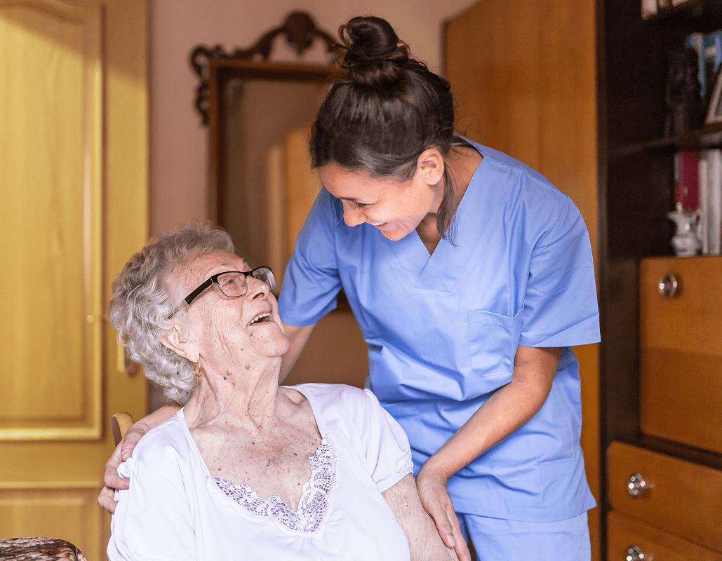 A nurse in blue scrubs is smiling and leaning toward an elderly woman with gray hair and glasses, who is sitting and smiling back. The nurse has one hand on the woman's shoulder and the other on her arm, engaging warmly with her. They are in a room with wooden furniture and a mirror in the background.