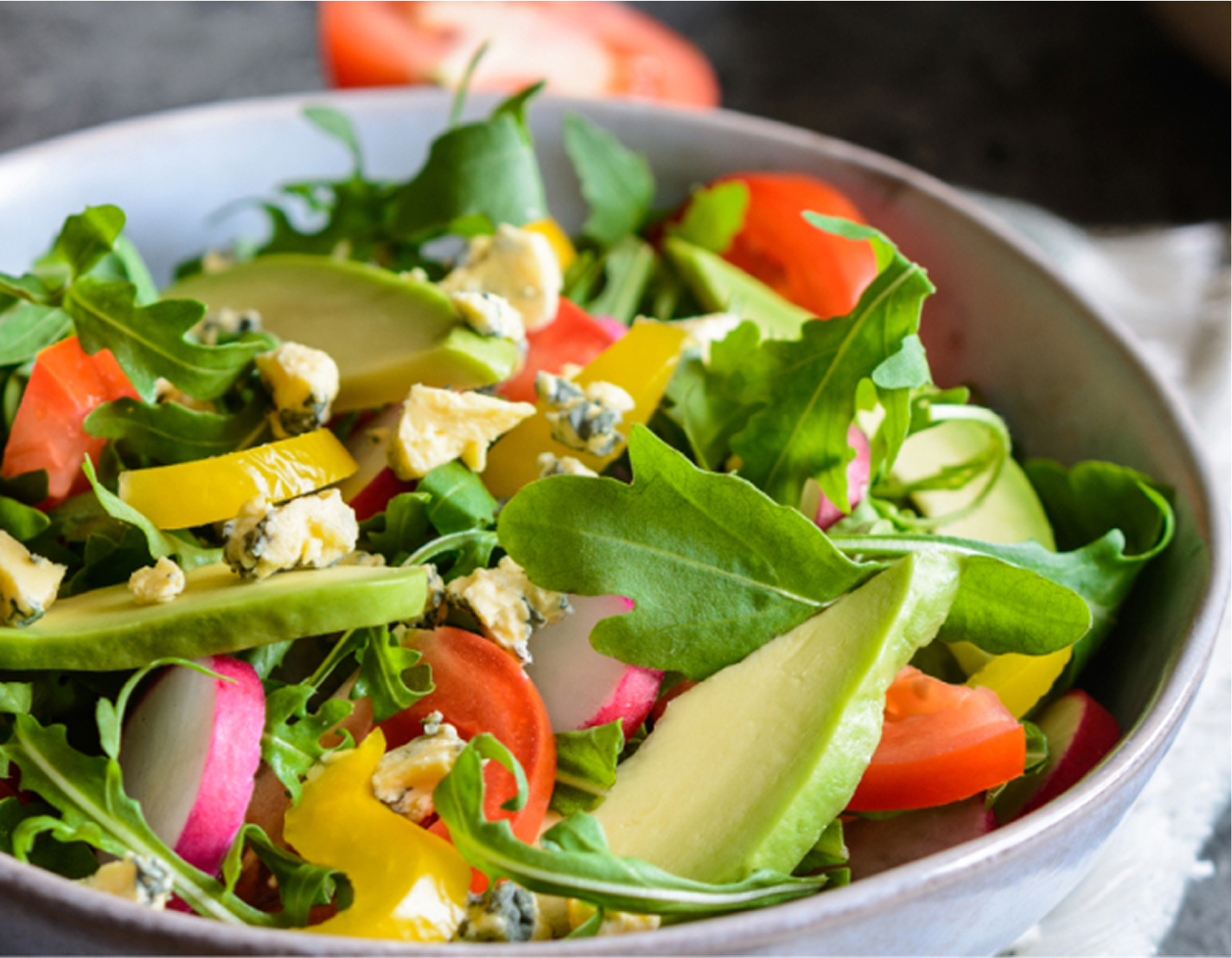A bowl of fresh salad containing arugula, sliced tomatoes, cucumber, yellow bell peppers, radishes, crumbled blue cheese, and avocado slices.