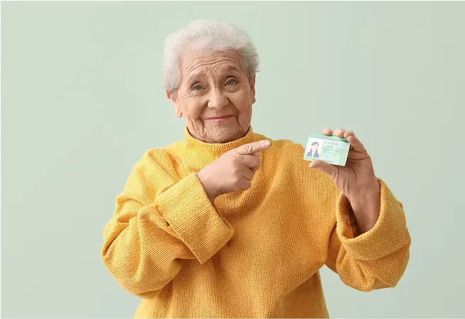 An elderly woman with gray hair, wearing a yellow sweater, smiling and pointing at a driver's license she is holding in her right hand against a light green background.