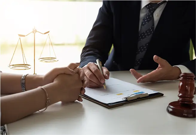 A person in a dark suit and polka dot tie is sitting at a table, signing a document, while another person with hands clasped on the table listens. A gavel and a set of scales are visible in the background, suggesting a legal or formal setting.
