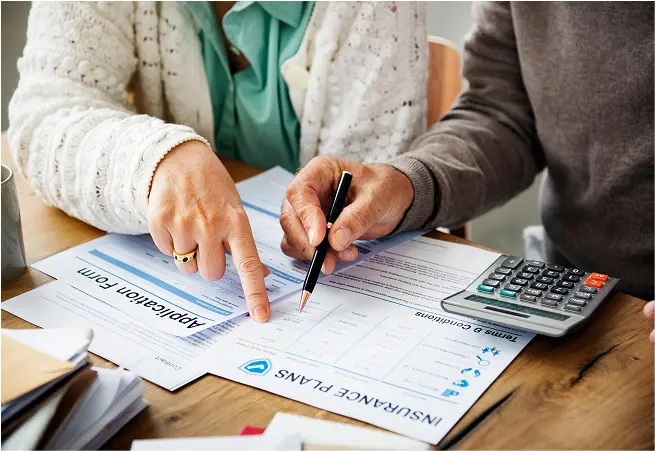 A person pointing at an insurance form with a pen, while another person holds a pen, on a wooden table. There is a calculator, a stack of papers, and a folder nearby.