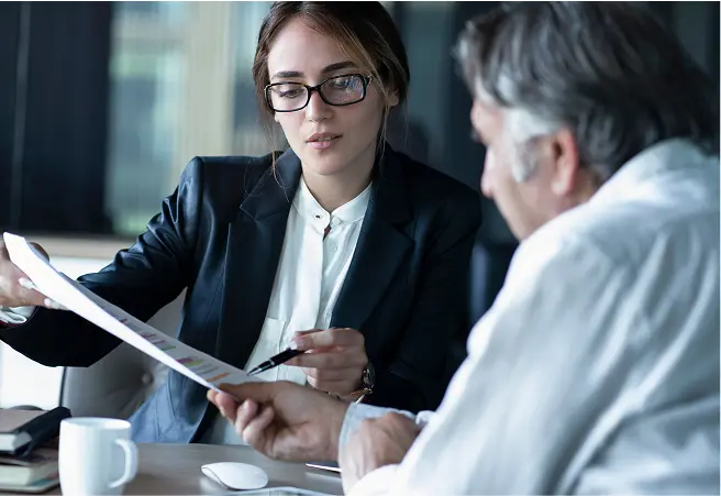 A young woman in professional attire, wearing glasses, and holding a document, discusses information with an older man with gray hair, who is also holding the document, in a well-lit office setting.