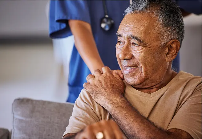 A senior man smiling while sitting on a couch, with a healthcare worker in blue scrubs standing behind him. The man appears content and comfortable.