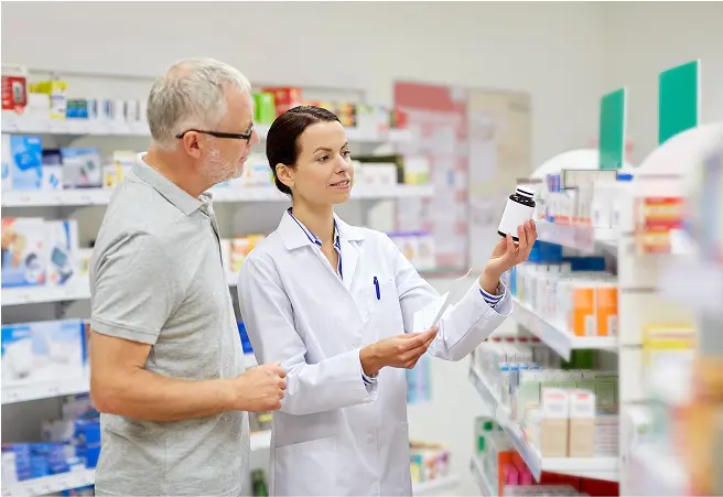 A female pharmacist in a white coat showing a medicine bottle to an older male customer in a pharmacy aisle with shelves stocked with various medication boxes.