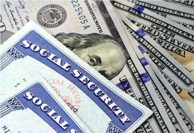 A close-up view of Social Security cards placed on top of various U.S. dollar bills, including $100 and $20 notes.