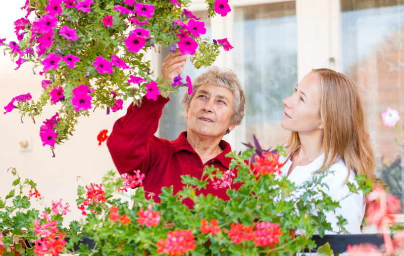 An elderly woman in a red sweater and a young woman with long blonde hair are standing among colorful flowering plants, with the elderly woman reaching up to touch or adjust pink flowers on a hanging basket. They are outdoors near a building with large windows.